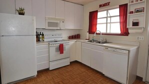 A compact kitchen with white cabinets, a microwave, stove, and dishwasher. A red towel hangs on the oven, and a window with red curtains is above the sink.