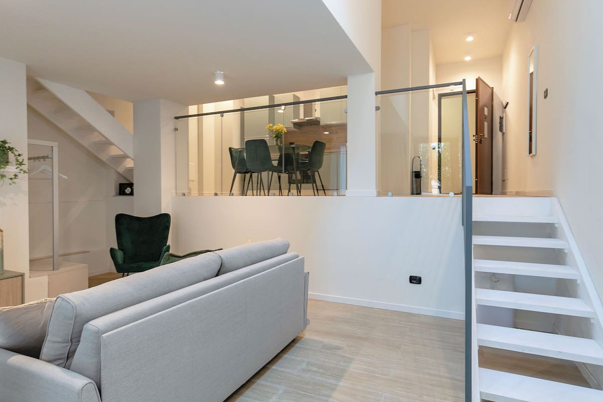 A modern living room with a grey sofa, a dining area with chairs and a table, and a staircase.
