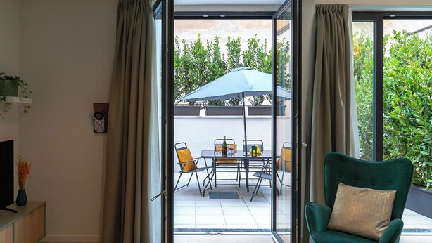 A room with a sliding glass door leading to an outdoor patio area with a table, chairs, and an umbrella. There is a green armchair with a cushion in the room.
