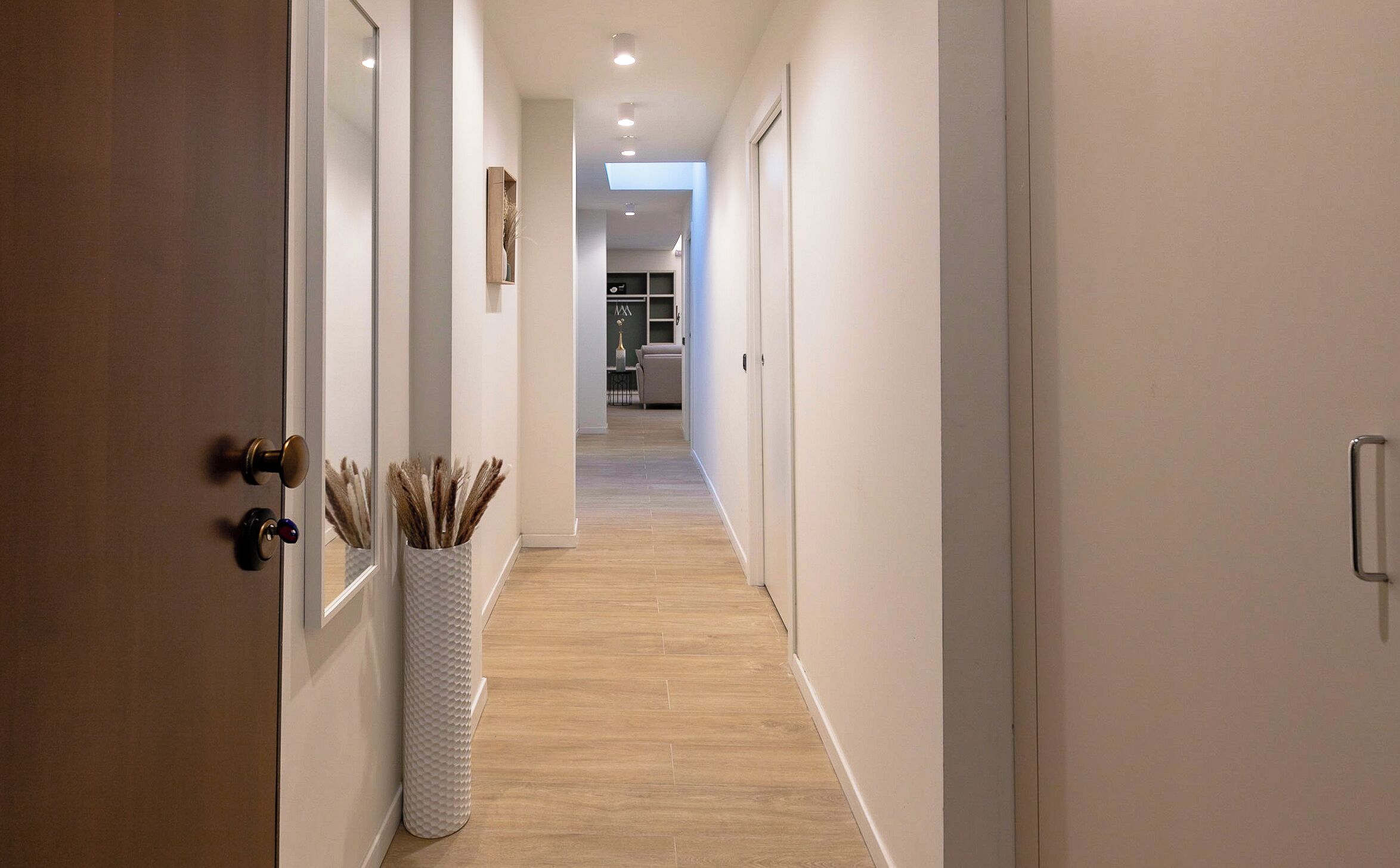 A hallway with a wooden floor, white walls, and a white vase with dried plants.