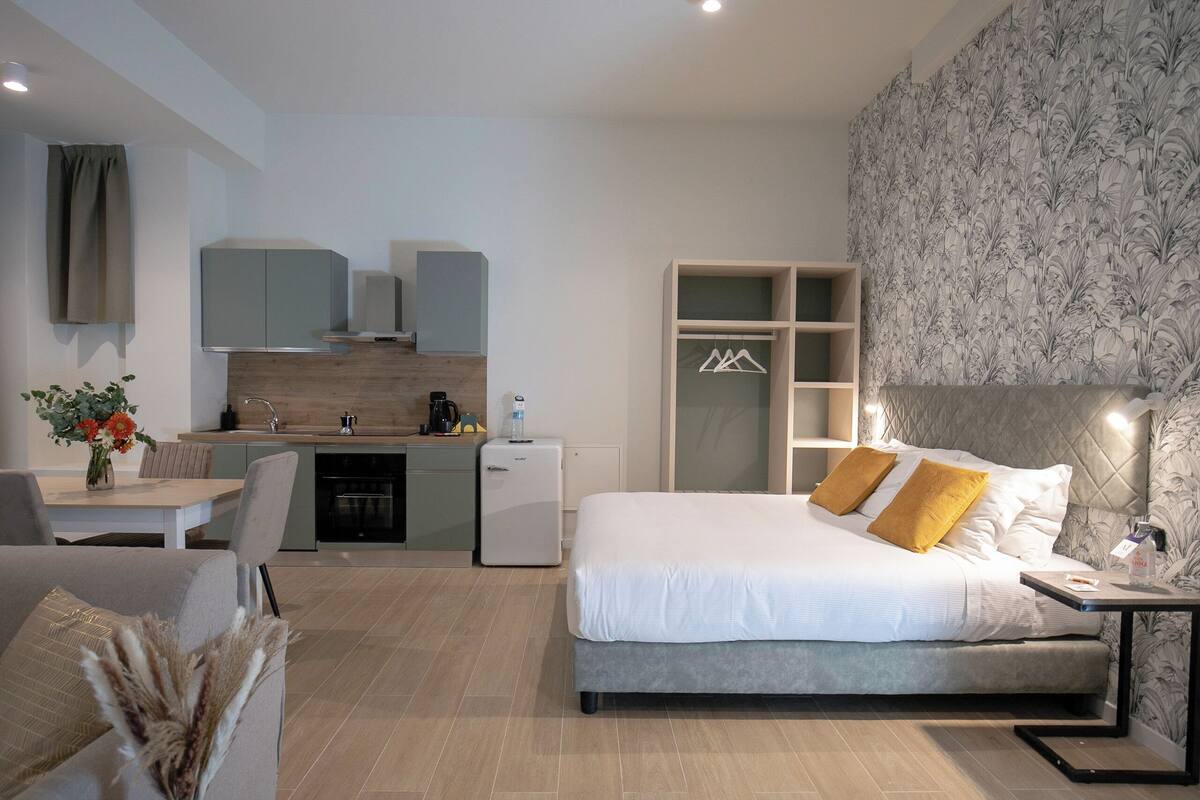 A modern hotel room with a large bed, a desk, a kitchenette, and a wardrobe.