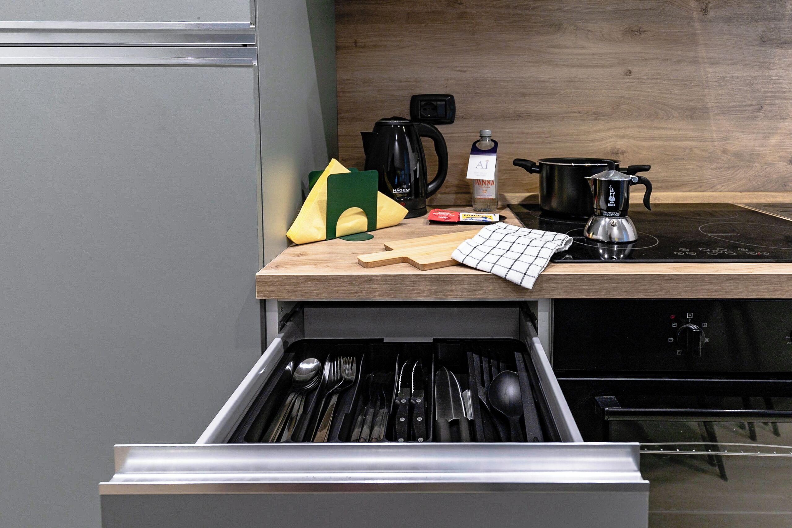 A kitchen with a drawer containing utensils, a cutting board, a kettle, a pot, and a milk carton.