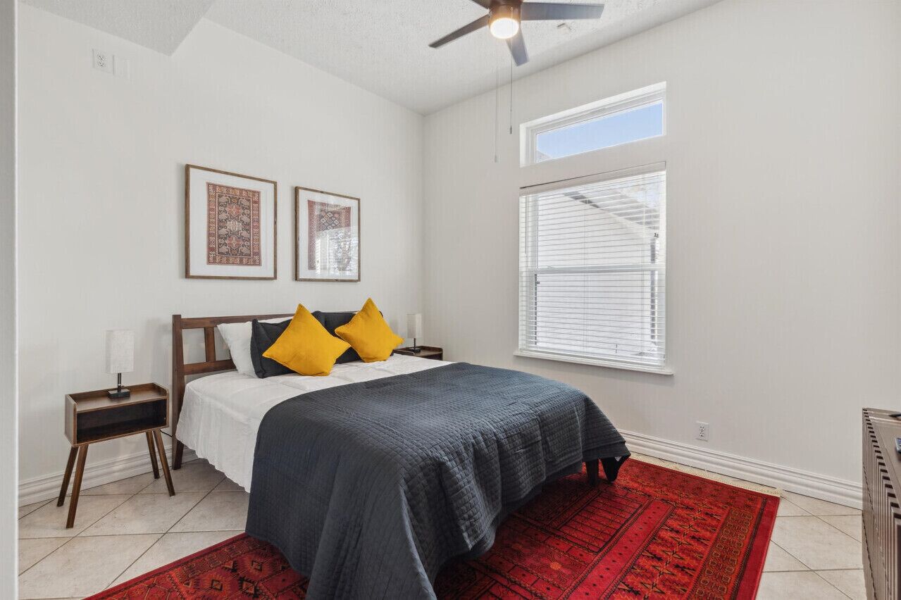 A bedroom with a bed, two nightstands, a ceiling fan, a window with blinds, and a red rug.