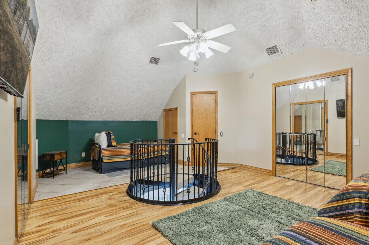 A bedroom with a wooden floor, a bed with a striped bedspread, a ceiling fan, a door, and a mirror.