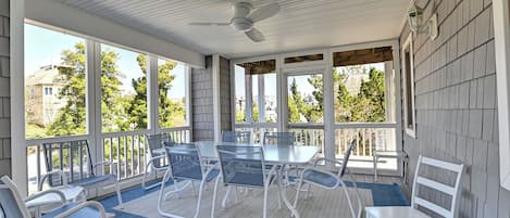 A covered patio with a table and chairs, a ceiling fan, and large windows offering views of the neighborhood.