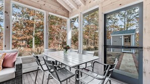 A covered patio with a wooden ceiling, a dining table with chairs, and a view of a house through large glass doors.