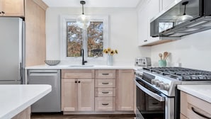 A modern kitchen with wooden cabinets, a built-in dishwasher, a gas stove, and a window with a view of trees.