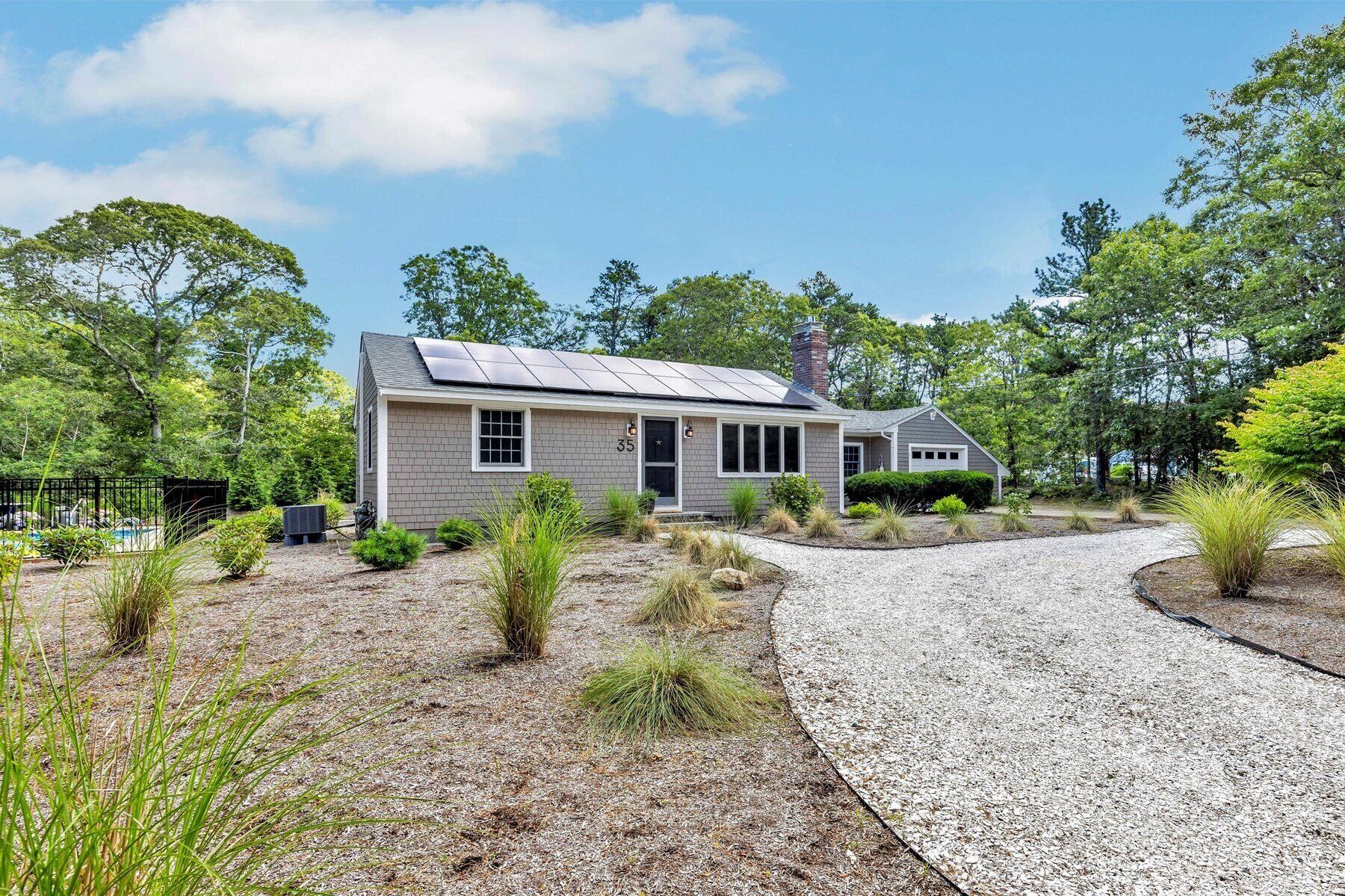A single-story house with a grey exterior, solar panels on the roof, and a gravel driveway leading to the entrance.
