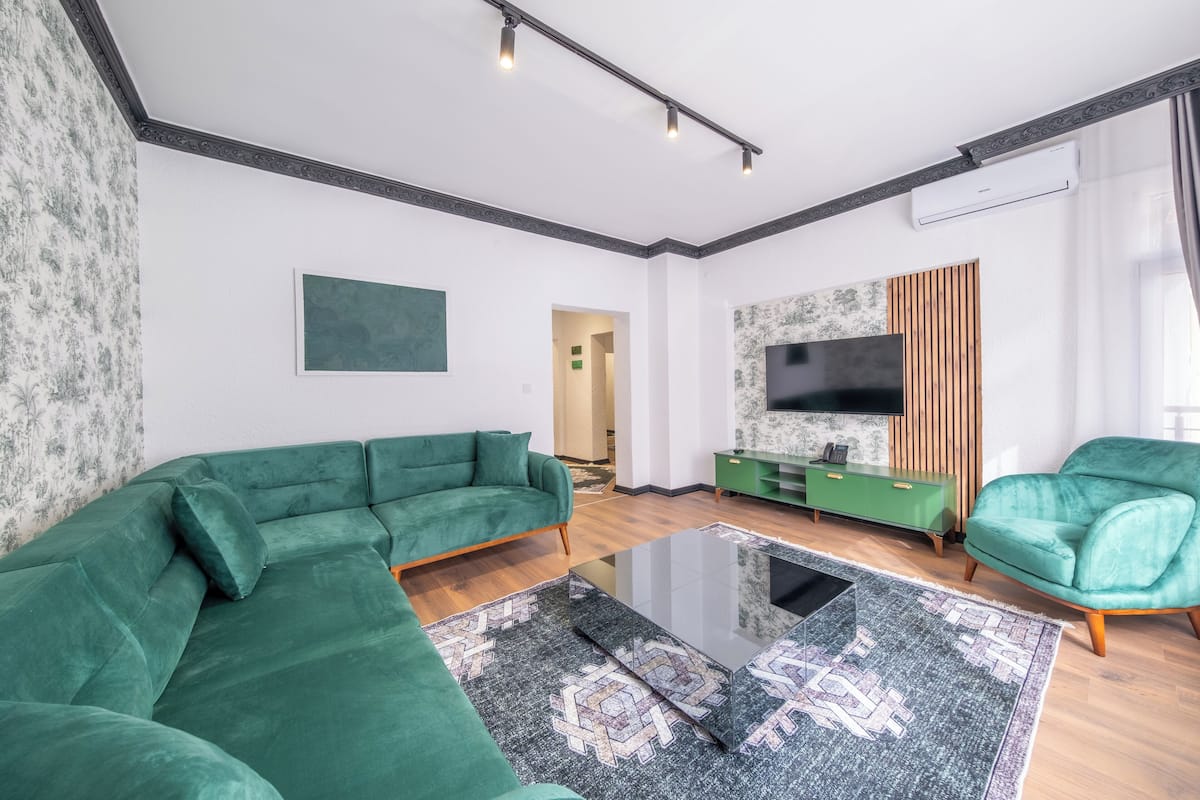 A modern living room with a green sofa, a glass coffee table, and a flat-screen TV mounted on the wall.