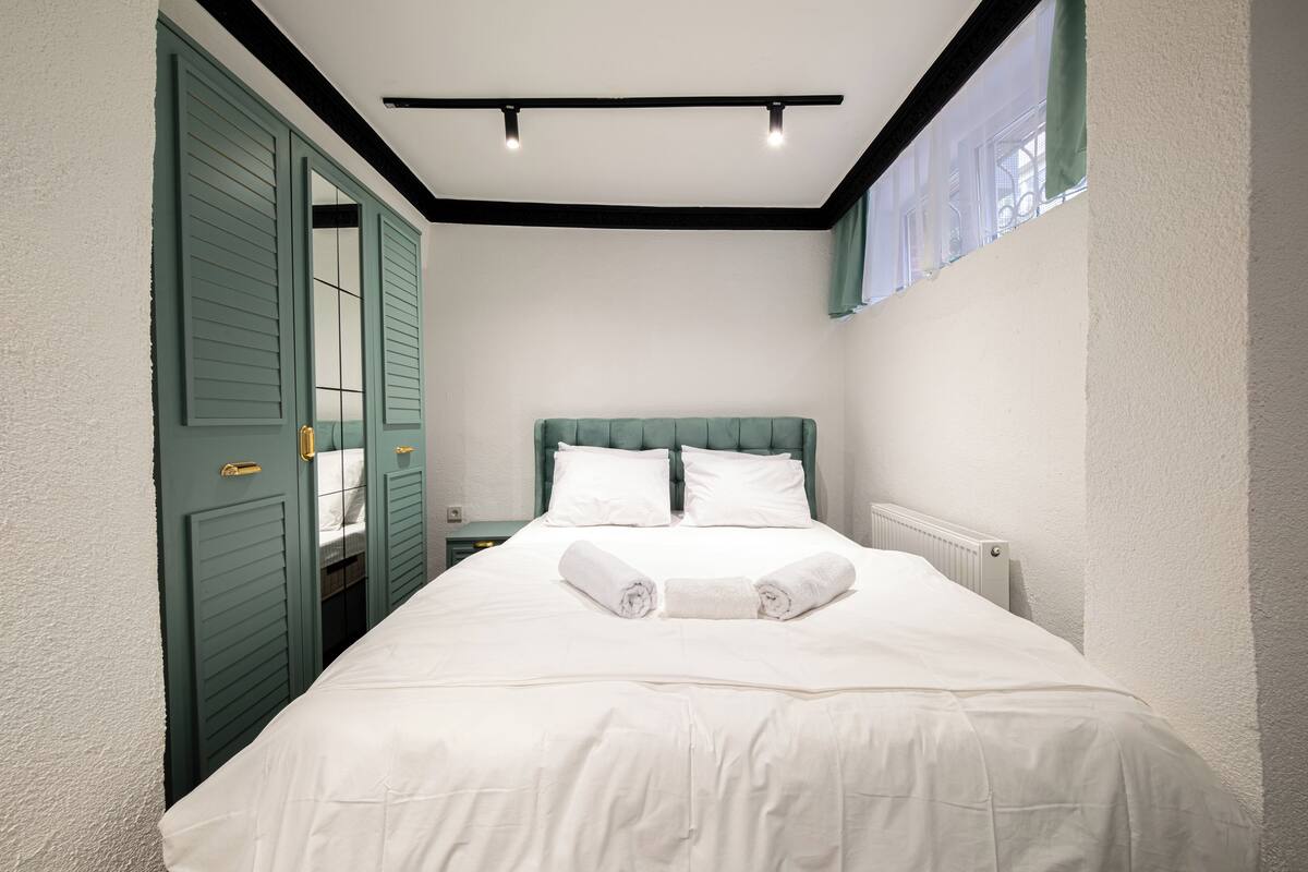 A neatly made bed with white linens and two rolled towels, a green headboard, and a window with a view.