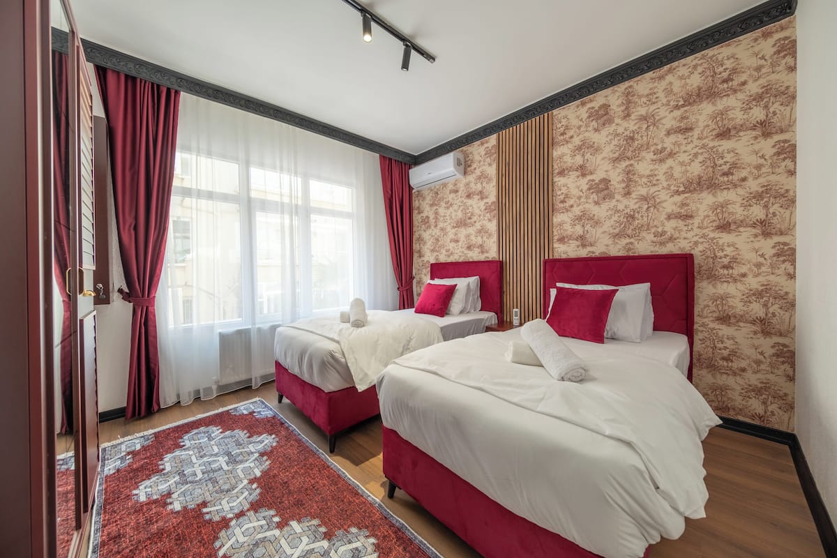A hotel room with two beds, a large window with curtains, a patterned wall, and a red and white rug.