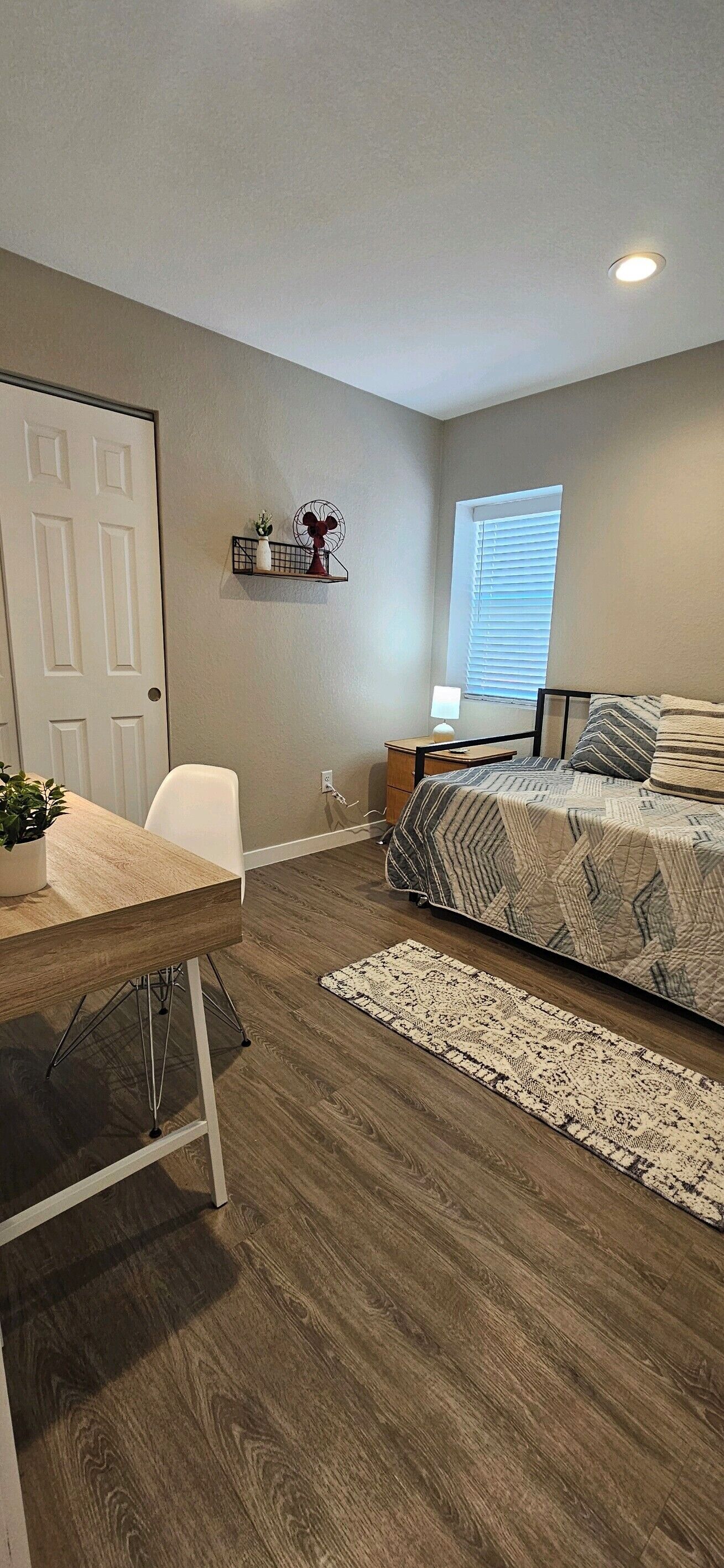 A bedroom with a bed, a desk, a chair, a window with blinds, a shelf with decorative items, and a door.