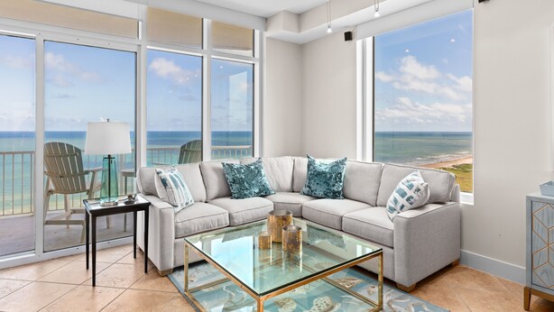 A spacious living room with a large glass coffee table, a beige sectional sofa with patterned cushions, and a view of the ocean through large windows. - Sapphire Condo & Spa Unit # 2106 3 Bedroom Condo (South Padre Island)