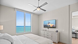 A bedroom with a large window, a ceiling fan, a TV on a white cabinet, and a bed with white bedding. - Sapphire Condo & Spa Unit # 2106 3 Bedroom Condo (South Padre Island)