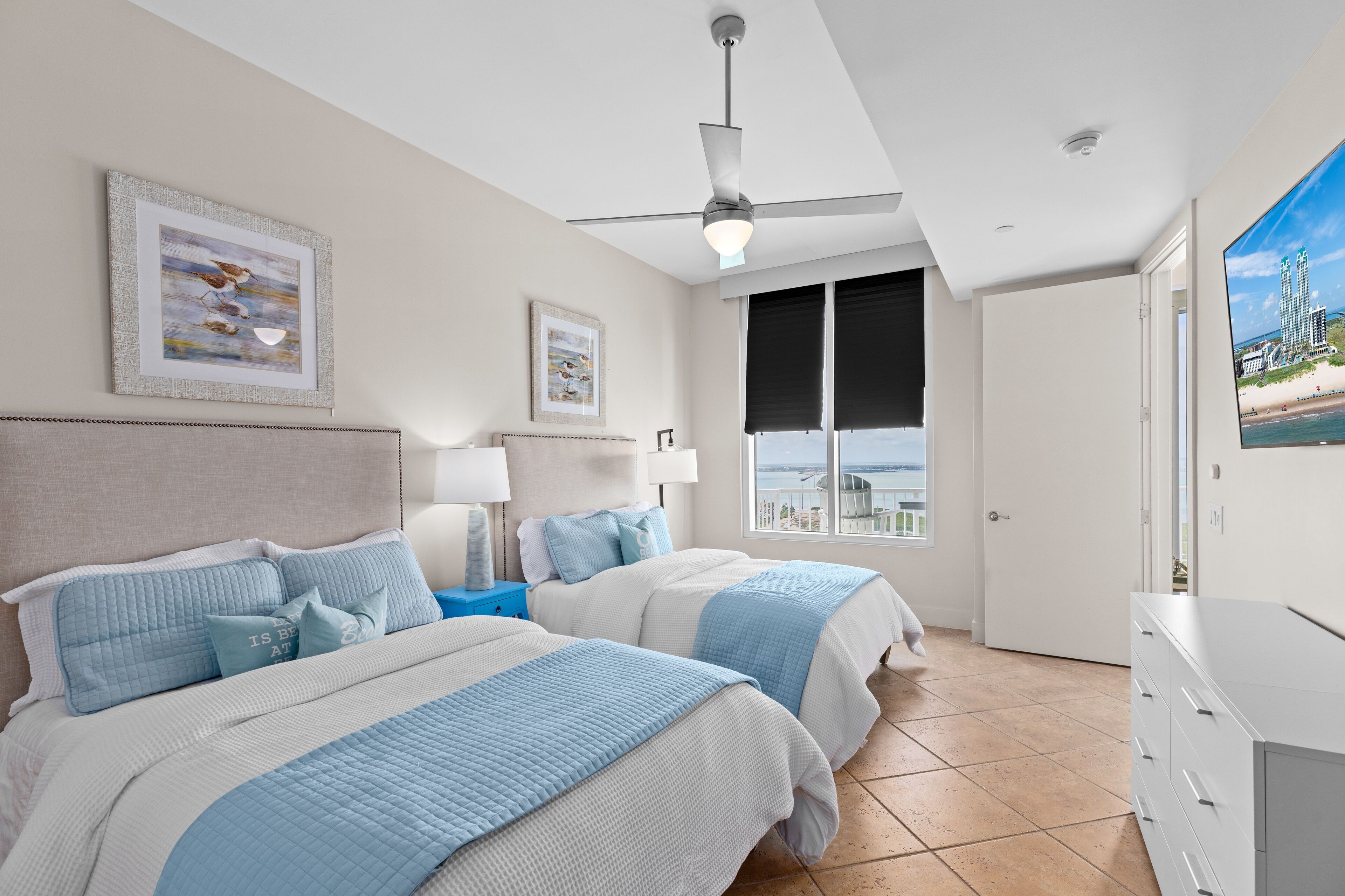 A bedroom with two beds, a ceiling fan, a nightstand, a dresser, and a large window with a view of a beach and buildings.