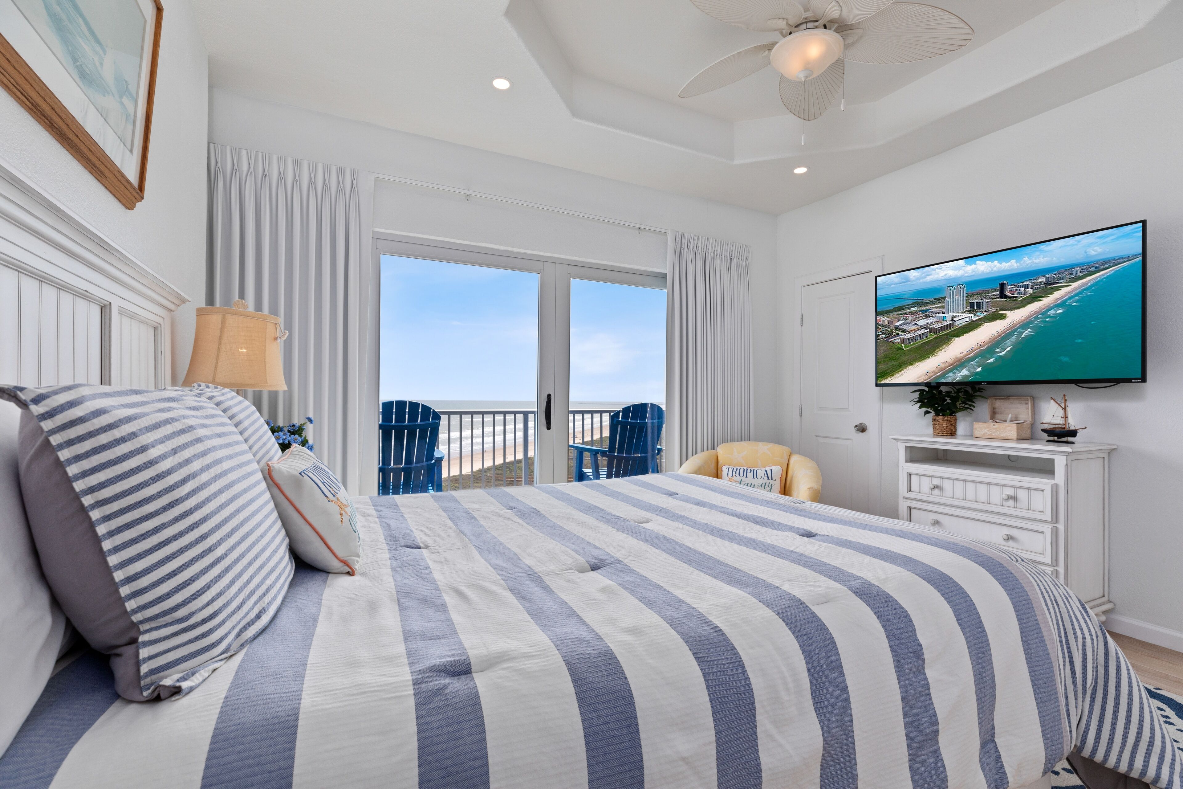 Habitación de hotel con una cama grande, un televisor que muestra una vista de la playa, un balcón con vistas al océano y un ventilador de techo.
