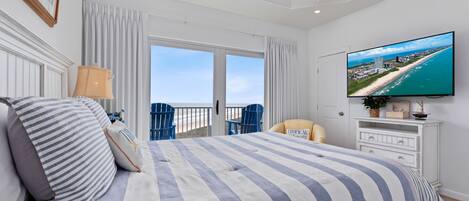 Habitación de hotel con una cama grande, un televisor que muestra una vista de la playa, un balcón con vistas al océano y un ventilador de techo.
