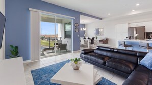 Een moderne woonkamer met een zwarte leren bank, een witte salontafel en een blauw vloerkleed. Er is een schuifdeur naar een balkon met uitzicht op een woonwijk.