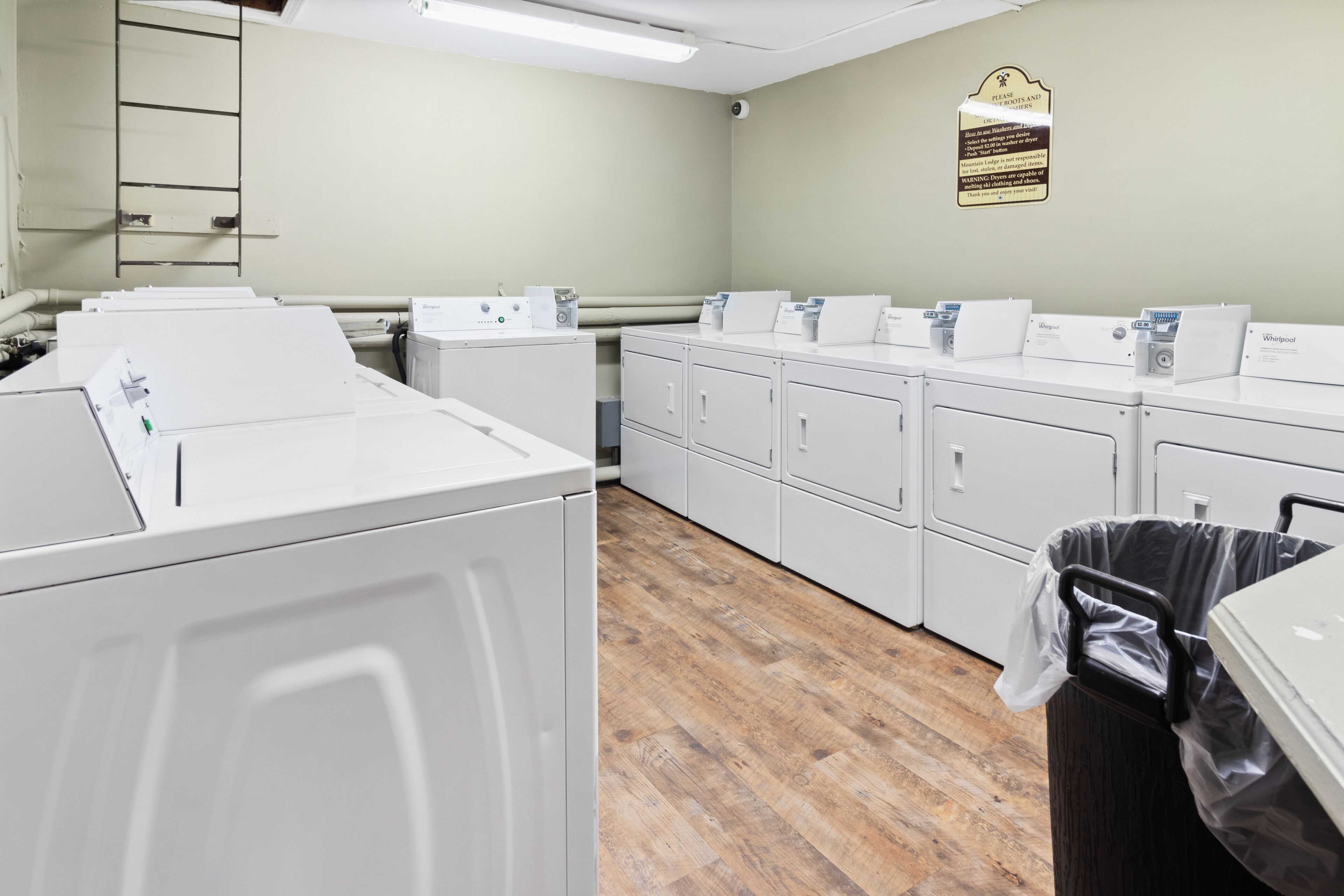 A laundry room with multiple white washers and dryers, wooden flooring, and a sign on the wall.
