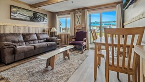 Ein gemütliches Wohnzimmer mit einer braunen Ledersofa, einem Holztisch und einem Esstisch mit Holzstühlen. Ein großes Fenster bietet Blick auf ein Gebäude draußen.