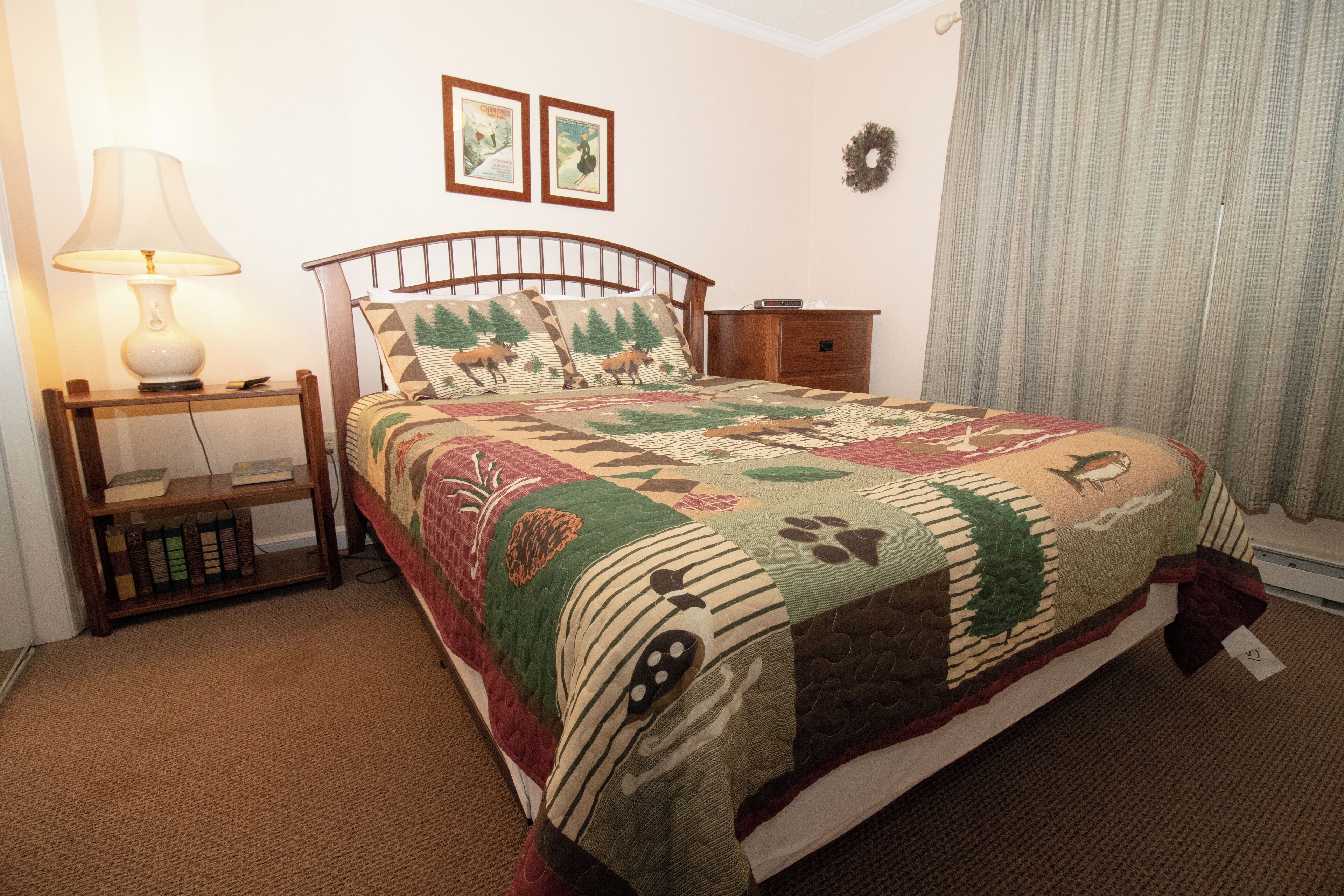 A neatly made bed with a quilt, a bedside table with a lamp, and a small shelf with books.