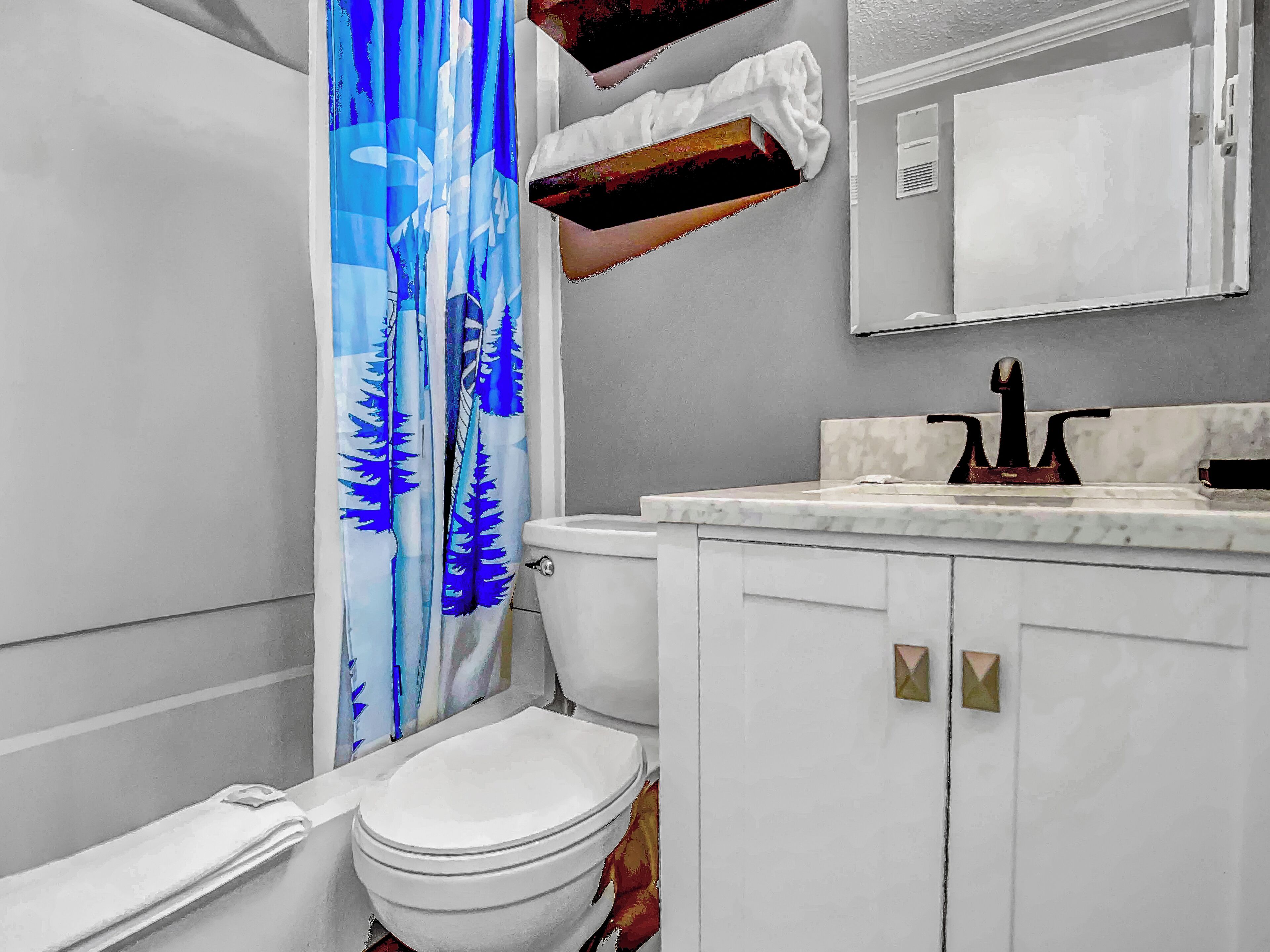 Un baño con inodoro, lavamanos con espejo y cortina de ducha con estampado de bosque azul.