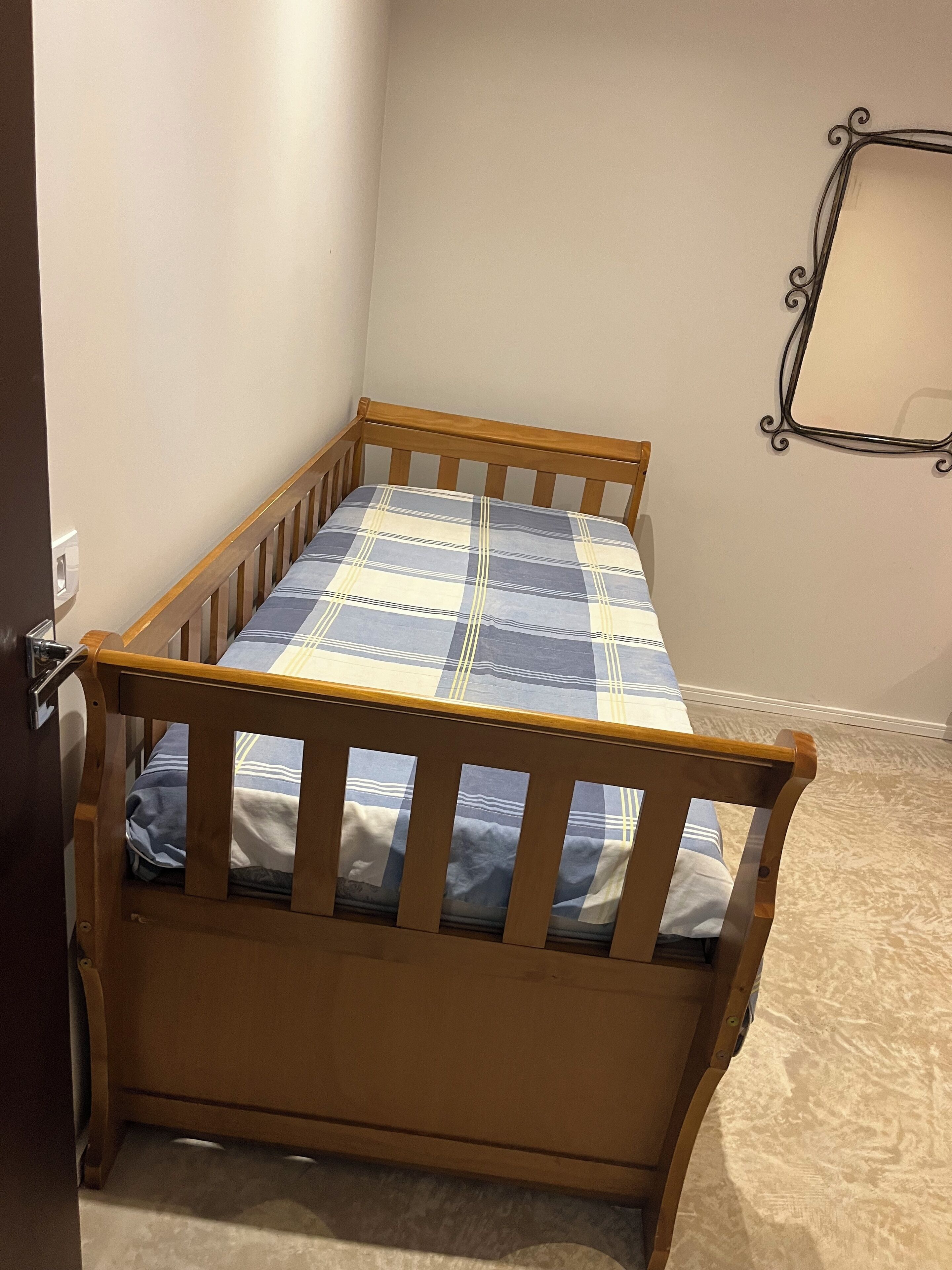A small, wooden bed with a plaid bedspread in a corner room with a plain wall and a metal picture frame.