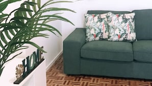 Une pièce avec un canapé vert, une grande plante en pot et deux éléments décoratifs suspendus au mur.