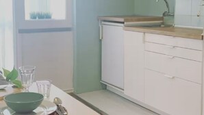 Une cuisine compacte avec des meubles blancs, un four à micro-ondes et un évier. On y trouve une table à manger avec un saladier et des cuillères, ainsi qu’un tapis à motifs sur le sol.