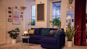 A living room with a dark blue sofa, a small side table with a lamp, and several potted plants.
