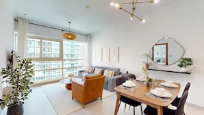 A modern living room with a sofa, dining table, and chairs. There is a large window with blinds, a hanging light fixture, and a mirror on the wall.