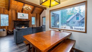 A cozy cabin interior with a fireplace, a large wooden dining table, and a view of a cabin through a large window. - Hare 12 | Hot Tub | Dogs OK | Foosball + Darts | Bikes | 3BR/2BA (Sunriver)