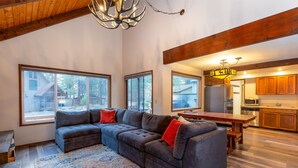 A spacious living room with a sectional sofa, large windows, a wooden ceiling, and a kitchen area. - Hare 12 | Hot Tub | Dogs OK | Foosball + Darts | Bikes | 3BR/2BA (Sunriver)