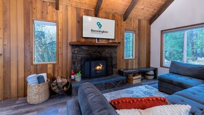 A cozy living room with a stone fireplace, a large flat-screen TV mounted above it, and a comfortable seating area with sofas and a rug. - Hare 12 | Hot Tub | Dogs OK | Foosball + Darts | Bikes | 3BR/2BA (Sunriver)