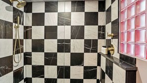 A bathroom with black and white checkered marble tiles, a gold showerhead, and a pink-framed window.