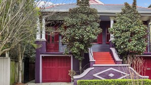 A two-story house with a red garage door, a staircase leading to the second floor, and a small front yard with a tree and shrubbery.
