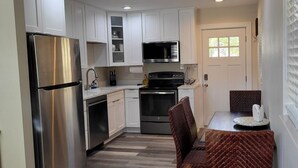 A compact kitchen with white cabinets, a stainless steel refrigerator, a microwave, and a stove. There is a dining table with chairs and a window with blinds.