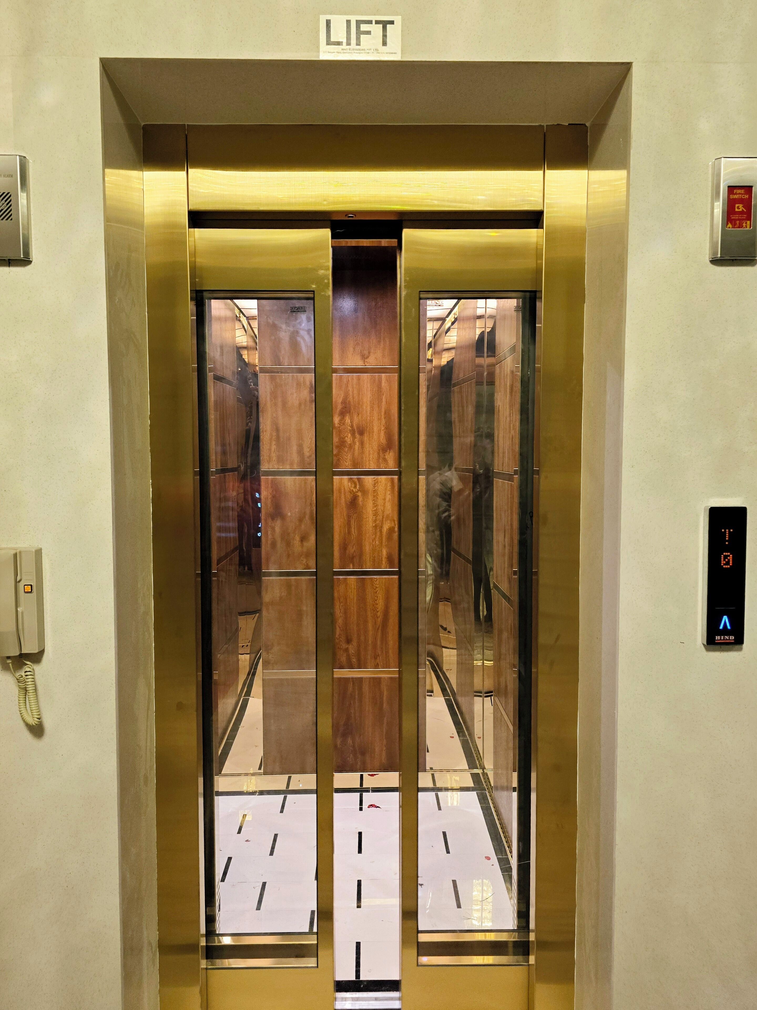 Un ascensor con acabado metálico, un cartel con la palabra 'LIFT' en la parte superior y un interruptor de incendios a la derecha.