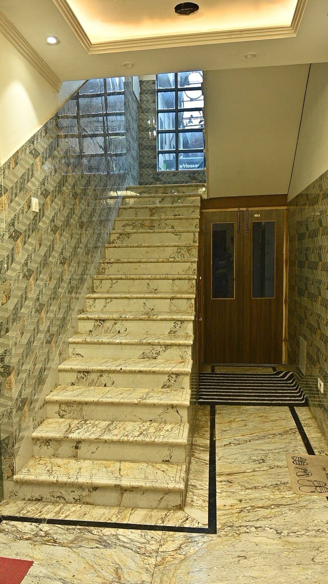Una escalera de mármol conduce a una puerta de madera con paneles de vidrio, en un espacio rodeado de paredes con diseños.