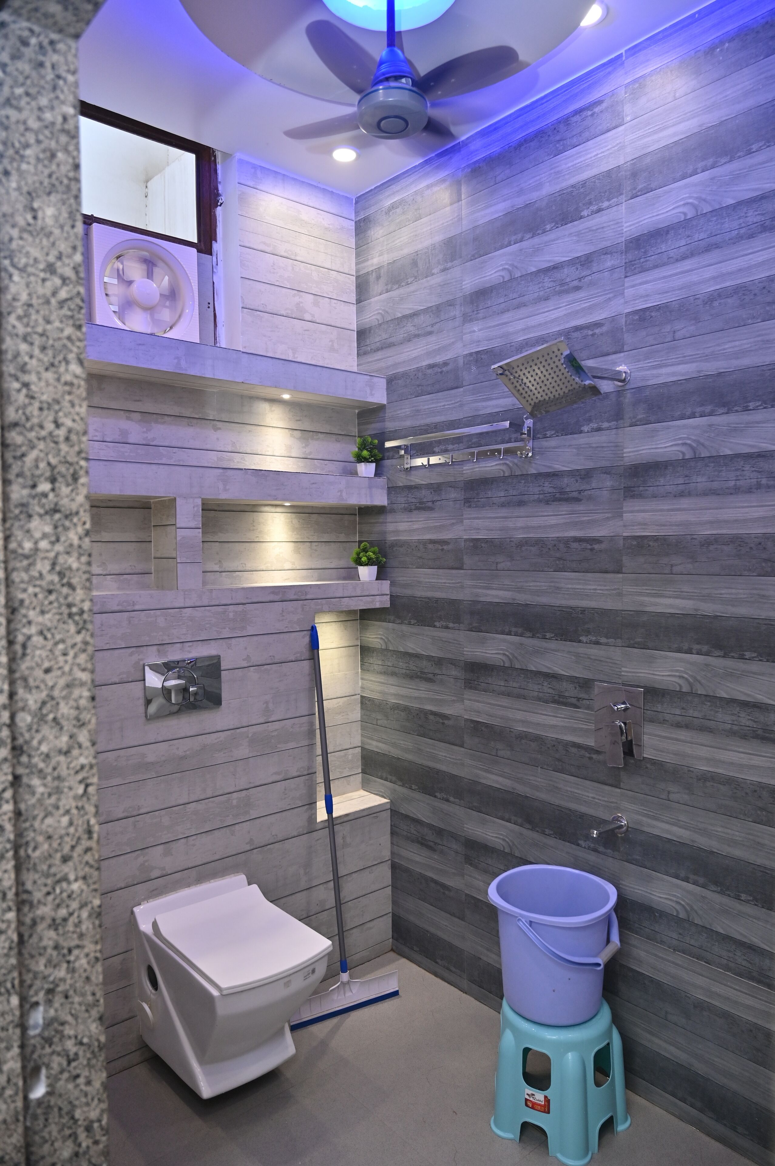 A modern bathroom with a grey tiled shower, a white toilet, a blue bucket on a stool, and a ceiling fan.