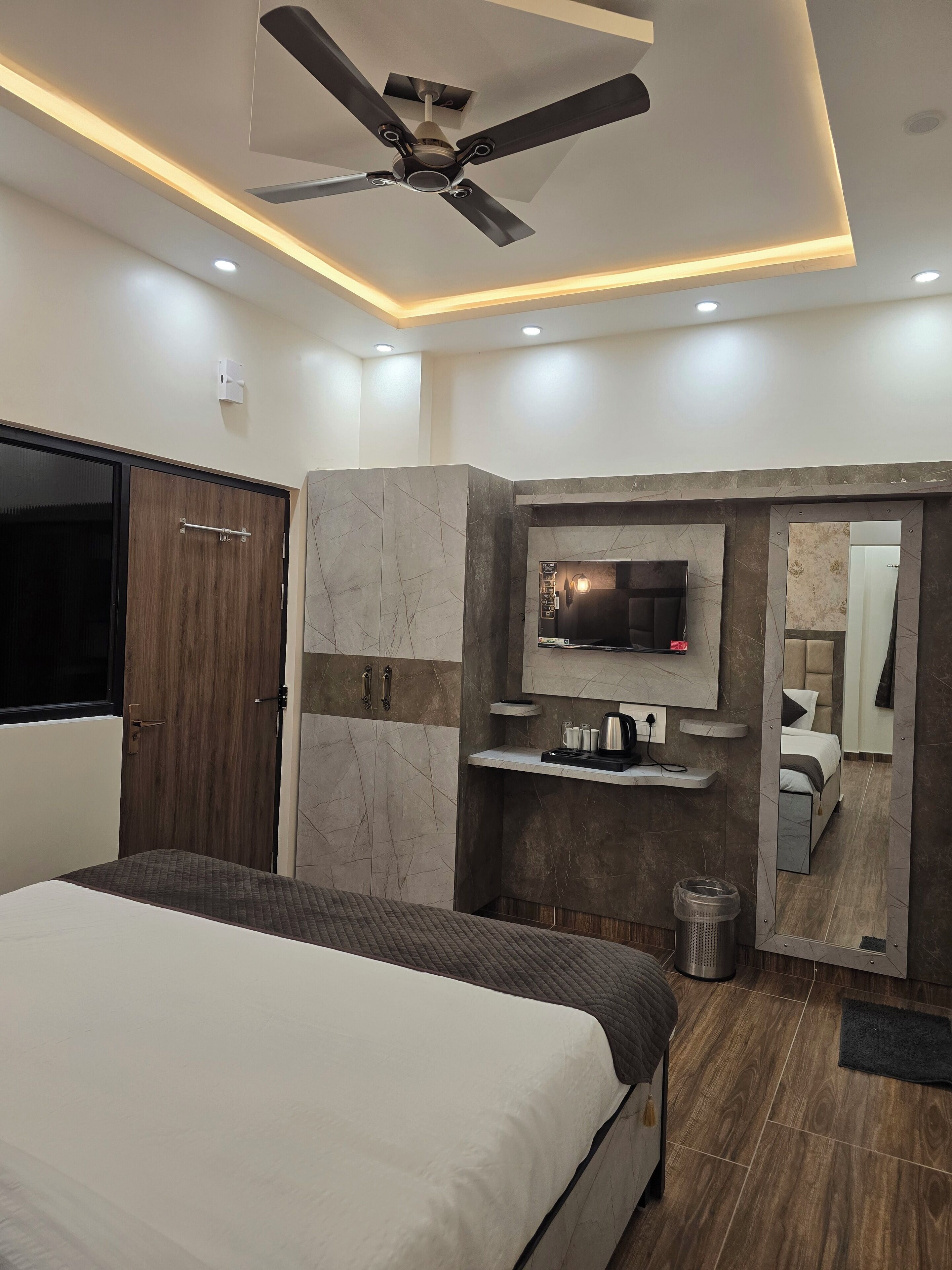 Ein modernes Schlafzimmer mit Bett, Deckenventilator, Fernseher an einer Marmorwand und einem Spiegel, der den Raum reflektiert.