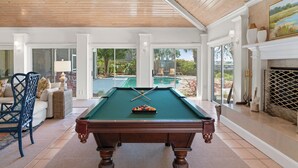 Una mesa de billar en una habitación bien iluminada con amplios ventanales, chimenea y vista a una piscina exterior.