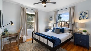 A bedroom with a bed, bedside tables, a desk, a chair, a ceiling fan, and a window with curtains.
