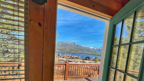 Una cabaña de madera con puerta y ventanas verdes, un balcón con vistas a montañas y un lago.