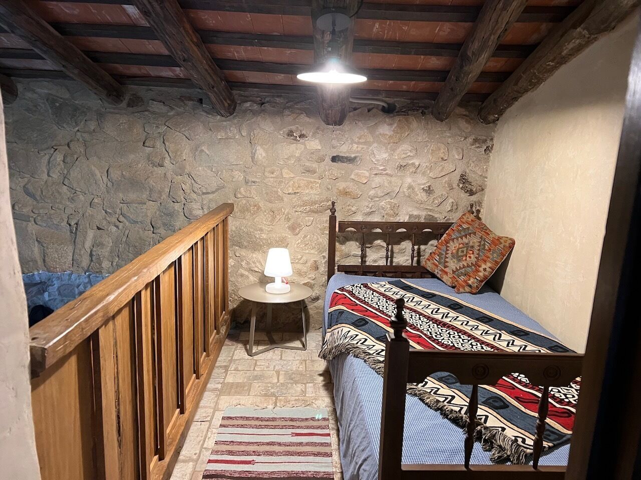 Une chambre avec un mur en pierre, des poutres en bois au plafond, un lit avec une literie à motifs, une table de chevet avec une lampe et un petit tapis.