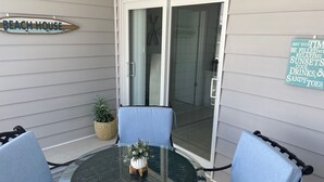 Een terras met een glazen tafel, twee blauwe stoelen, een bloempot en een bord met de tekst 'BEACH HOUSE'.