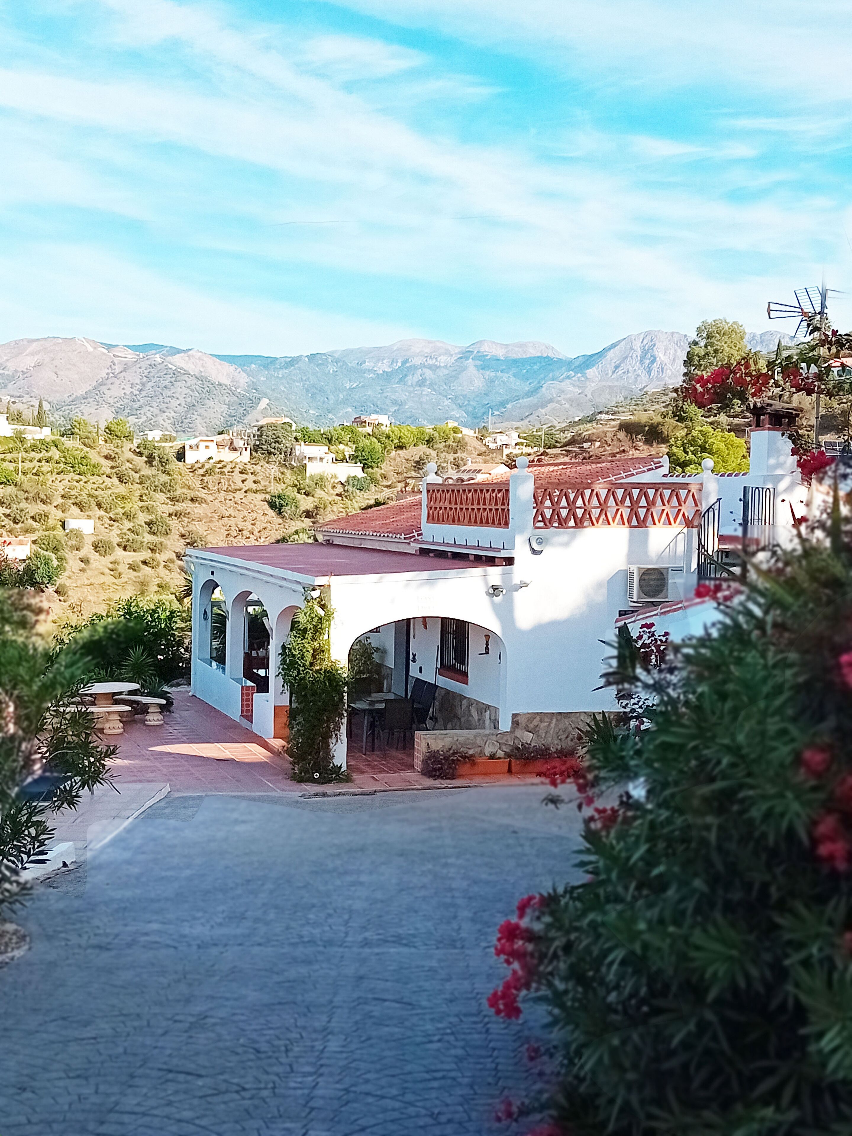Un edificio blanco con techo de tejas rojas y ventanas arqueadas, rodeado de vegetación y montañas al fondo.
