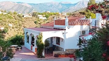 Un edificio blanco con techo de tejas rojas y ventanas arqueadas, rodeado de vegetación y montañas al fondo.
