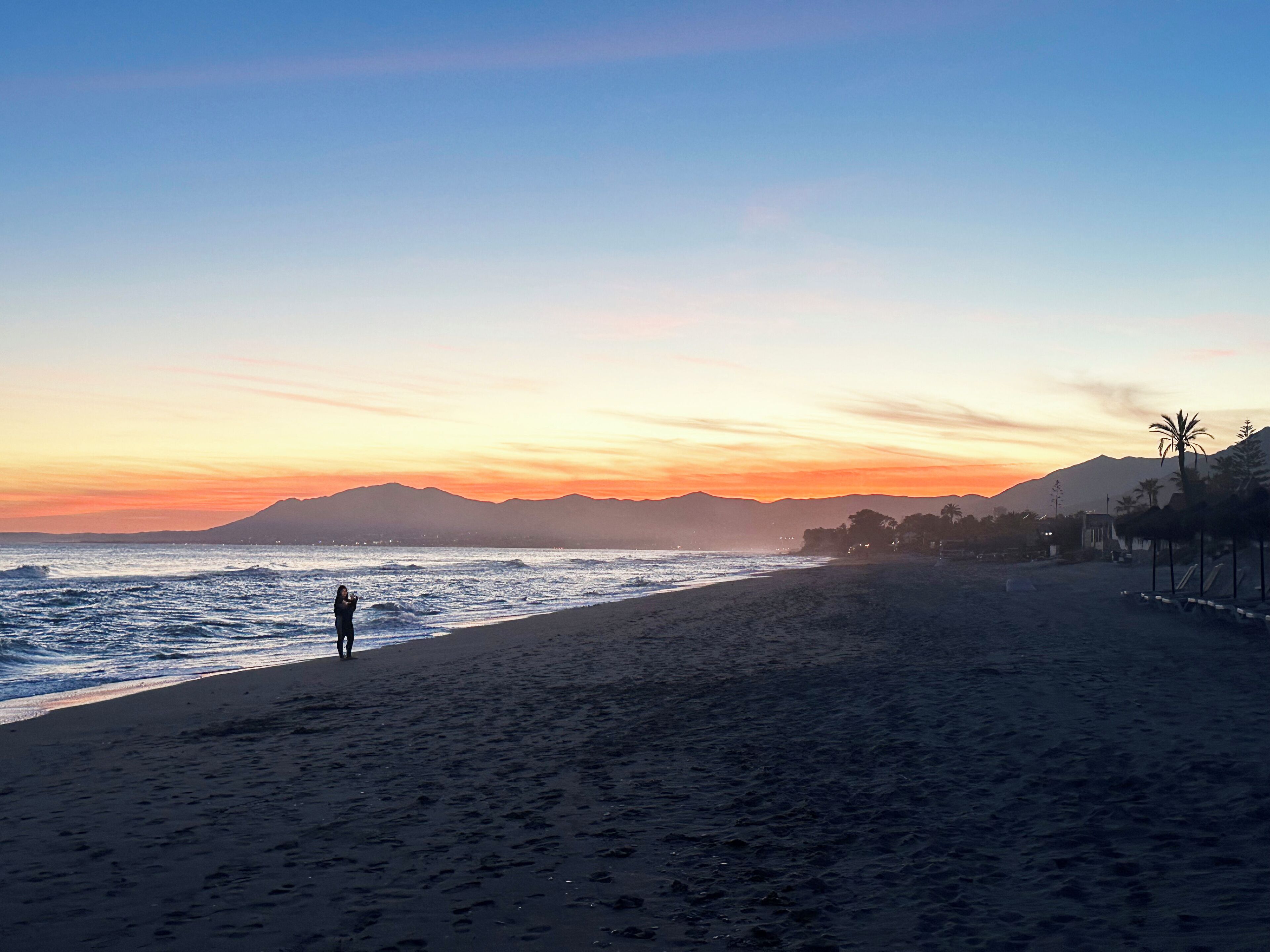 Una playa con una persona de pie cerca del agua, montañas al fondo y un cielo despejado.