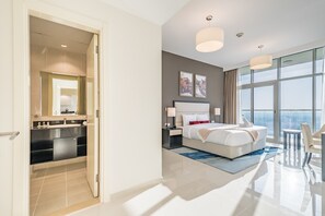 Habitación de hotel moderna con una cama grande, una zona de estar, un baño con tocador y vistas a la ciudad a través de amplias ventanas.
